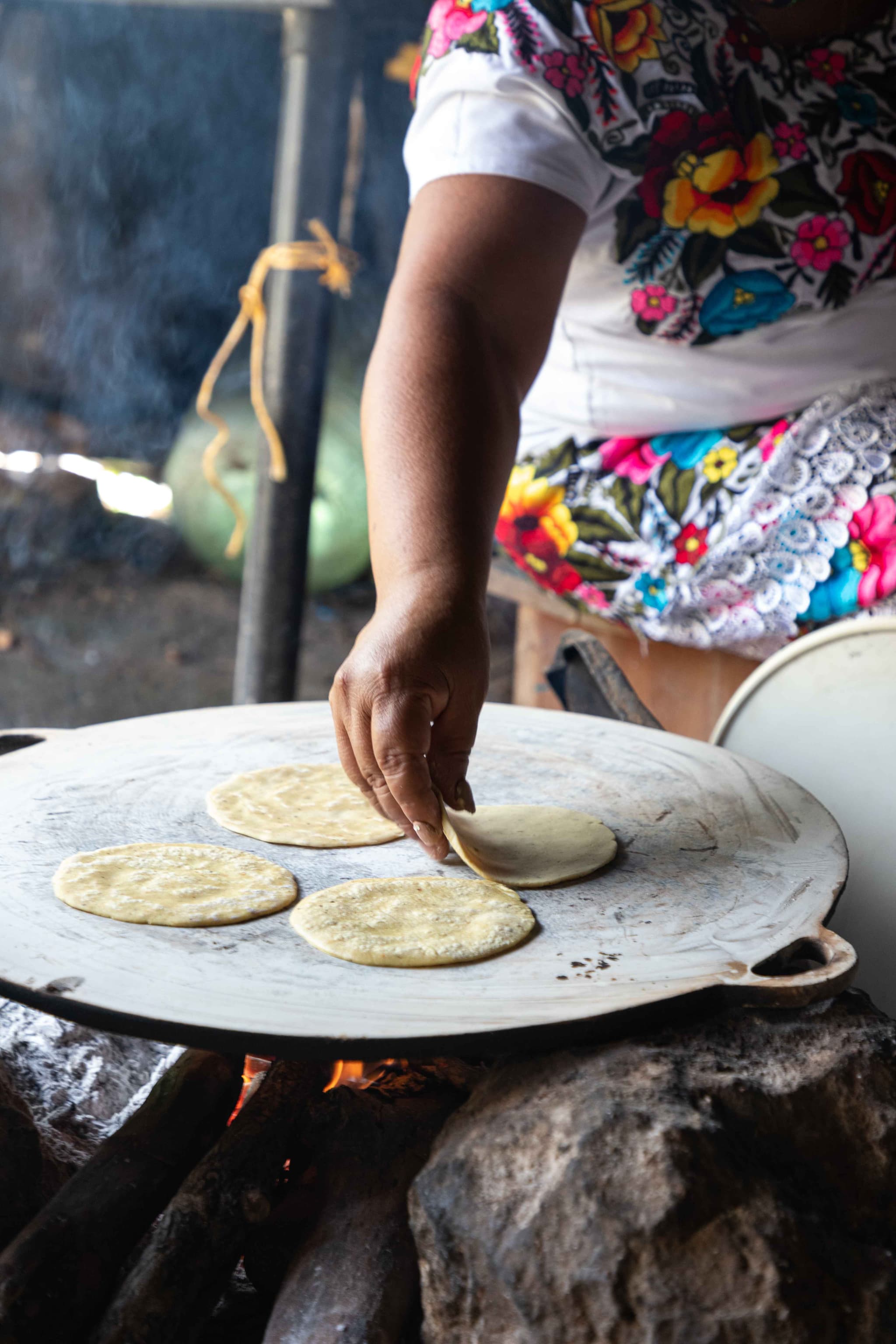 Cocinera con huipil bordado voltea tortillas de maíz en un comal sobre el fuego de leña